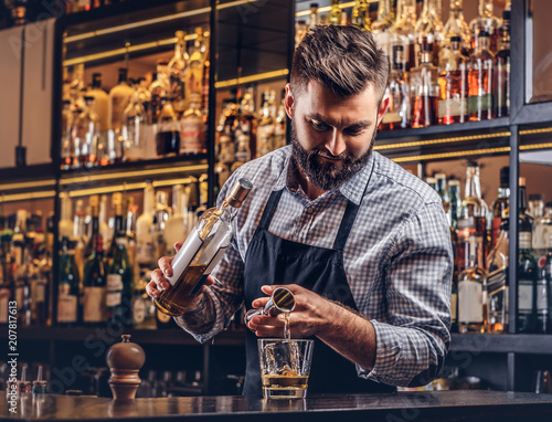 Stylish brutal barman in a shirt and apron makes a cocktail at bar counter background.