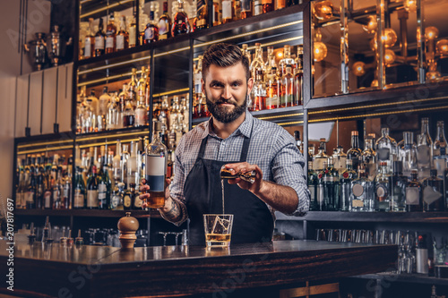 Stylish brutal barman in a shirt and apron makes a cocktail at bar counter background.