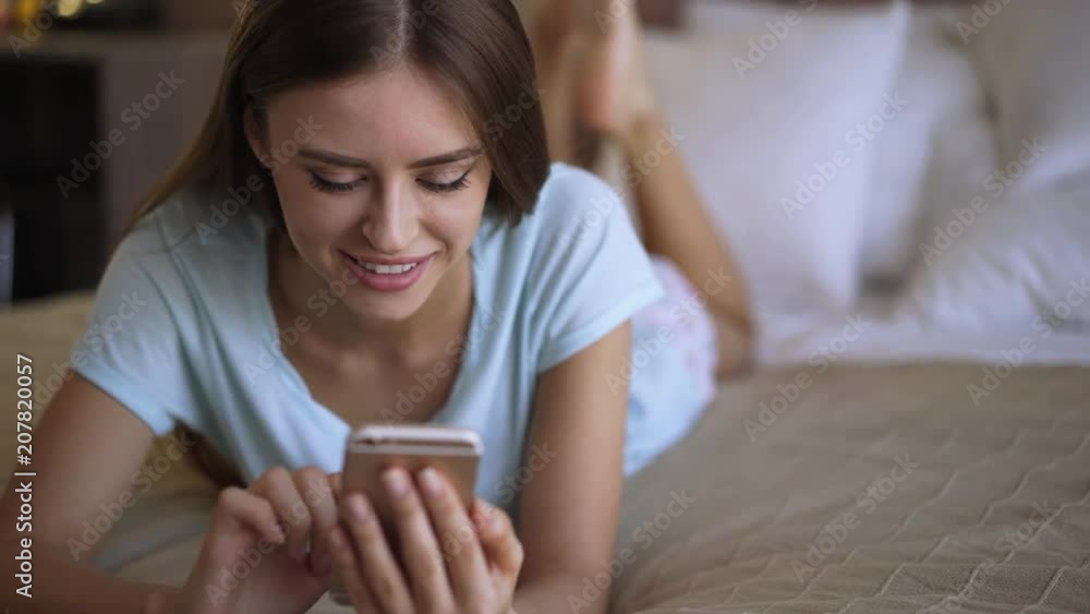 Attractive young woman wearing a white t shirt is smiling, typing on her smartphone and lying on bed. Slider real time medium shot Attractive young woman wearing a white t shirt is smiling, typing on her smartphone and lying on bed. Slider real time medium shot