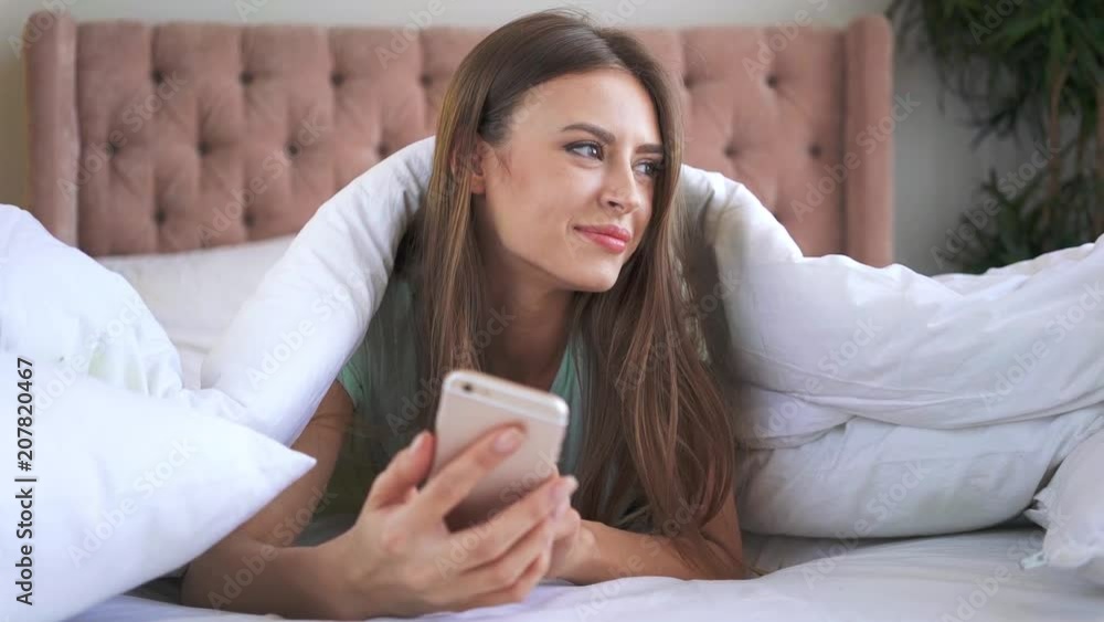 Attractive young woman with long hair wearing a night wear is lying under a blanket and texting. Handheld slow motion medium shot Attractive young woman with long hair wearing a night wear is lying under a blanket and texting. Handheld slow motion medium shot