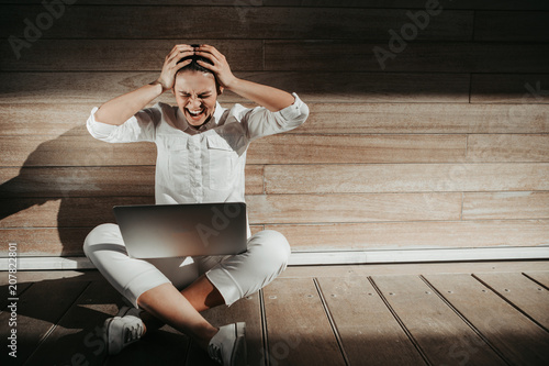 Portrait of upset woman sitting in timber room with laptop on her knees. She is holding her hands on head. Copy space in right side