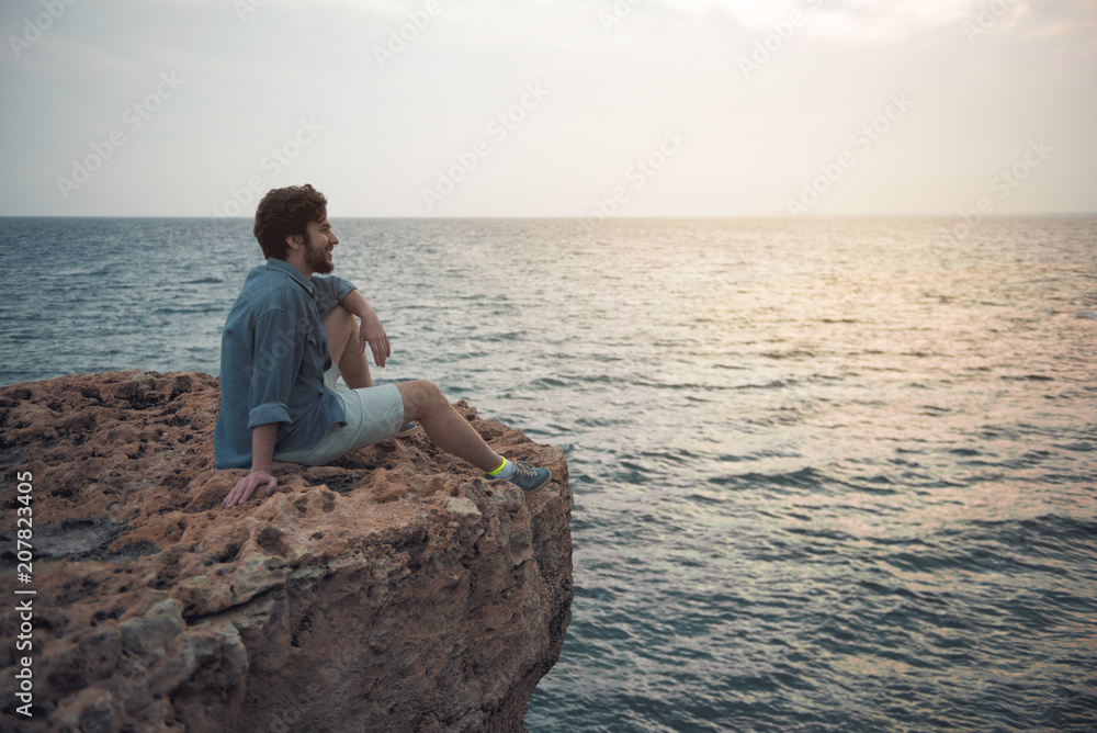 Full length side view profile of cheerful young bearded man is spending time outdoors while sitting on the edge of cliff. He is looking aside with slight smile. Copy space in the right side