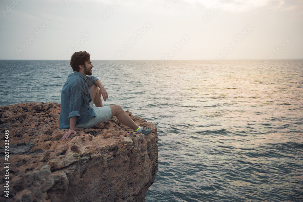 Full length side view profile of cheerful young bearded man is spending time outdoors while sitting on the edge of cliff. He is looking aside with slight smile. Copy space in the right side Full length side view profile of cheerful young bearded man is spending time outdoors while sitting on the edge of cliff. He is looking aside with slight smile. Copy space in the right side