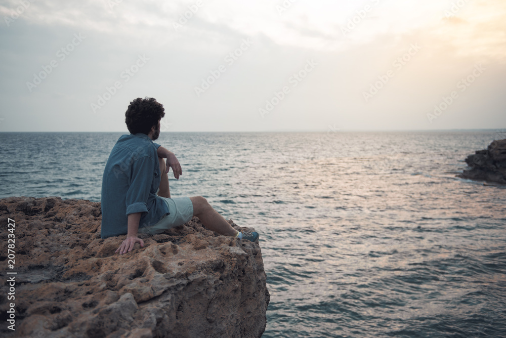 Charming dreamer. Full length back view of pensive stylish bearded male is sitting on the edge of cliff with endless ocean in background. He is looking at water thoughtfully. Copy space in right side Charming dreamer. Full length back view of pensive stylish bearded male is sitting on the edge of cliff with endless ocean in background. He is looking at water thoughtfully. Copy space in right side