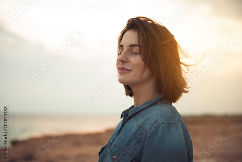 Great pleasure. Charming calm young woman is standing near sea with closed eyes and expressing delight. She is posing against wonderful sunset while enjoying last rays of the sun photo