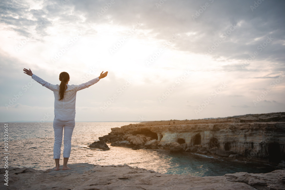 Happy life concept. Full length back view of charming girl is standing on rock with raised hands. She is looking at ocean with sunset in background. Copy space in the right side Happy life concept. Full length back view of charming girl is standing on rock with raised hands. She is looking at ocean with sunset in background. Copy space in the right side