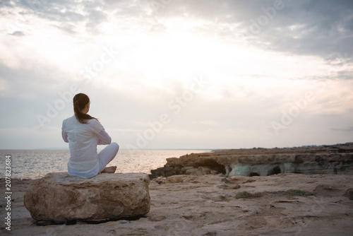 Inner harmony. Full length back view of calm girl is sitting on beach while meditating. She is practicing yoga on beach while enjoying wonderful view on the sea. Copy space in the right side