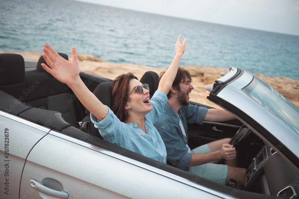 Full of joy. Overjoyed young romantic tourists couple is riding by luxury cabriolet along road with seascape in background. Woman is sitting with raised hands while enjoying summer vacation
