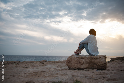 Wonderful view. Full length back view of thoughtful girl on beach is sitting on big round stone while enjoying sundown. She is looking at ocean and thinking dreamily. Copy space in the left side