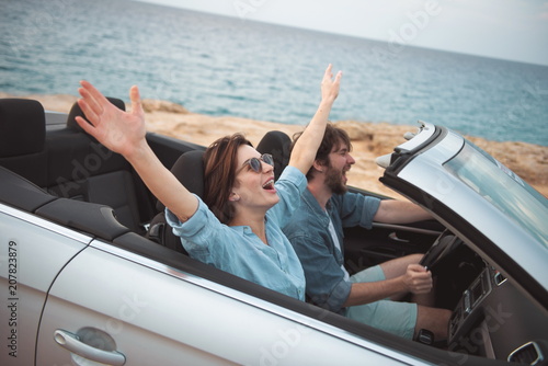 Full of joy. Overjoyed young romantic tourists couple is riding by luxury cabriolet along road with seascape in background. Woman is sitting with raised hands while enjoying summer vacation © Yakobchuk Olena