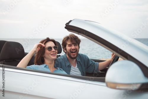 Happy time. Optimistic cute boyfriend and girlfriend are enjoying summer vacation. They are driving fashionable cabriolet with open roof along seaboard and expressing gladness. Romantic trip concept © Yakobchuk Olena