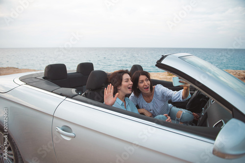 Selfie time. Young delighted women are sitting in cabriolet with open roof and taking picture while using modern mobile phone. They are waving and expressing gladness. Summer vacation concept