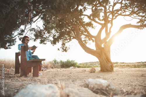 Full length of young attractive dreamy woman in sunglasses is sitting on bench and reading book with concentration. She is resting near big old tree against wonderful sunset. Copy space in right side