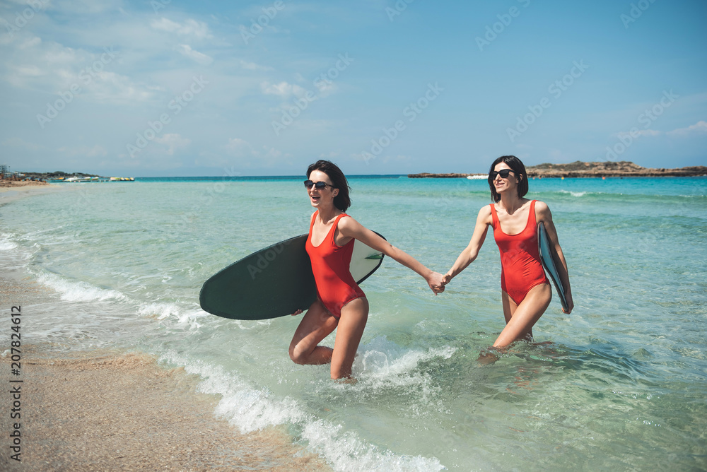 Full of joy. Full length of two gorgeous girls are holding hands while coming out of sea. They are carrying surfboards while expressing gladness and satisfaction after surfing. Summer vacation concept Full of joy. Full length of two gorgeous girls are holding hands while coming out of sea. They are carrying surfboards while expressing gladness and satisfaction after surfing. Summer vacation concept
