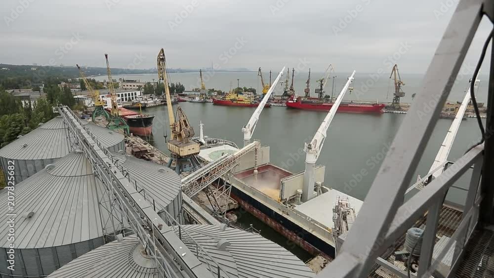 Panorama of ship loading grain crops on bulk freighter via trunk to open cargo holds at silo terminal in seaport. Cereals bulk transshipment to vessel. Transportation of agricultural products. Panorama of ship loading grain crops on bulk freighter via trunk to open cargo holds at silo terminal in seaport. Cereals bulk transshipment to vessel. Transportation of agricultural products.