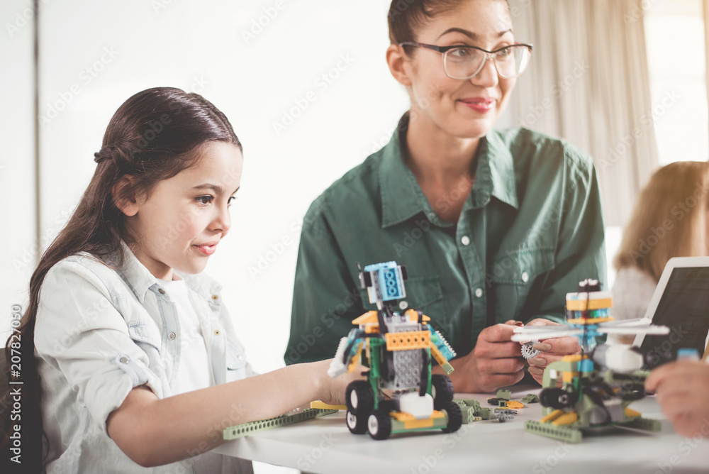 Happy child and glad teacher making robot from constructor while locating at desk. Contemporary technology concept