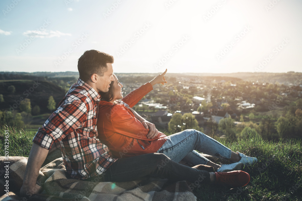 Look there. Full length side view of man and woman sitting and looking at beautiful summer sky. Delighted girl is pointing at shining sun. They are bonding to each other and smiling