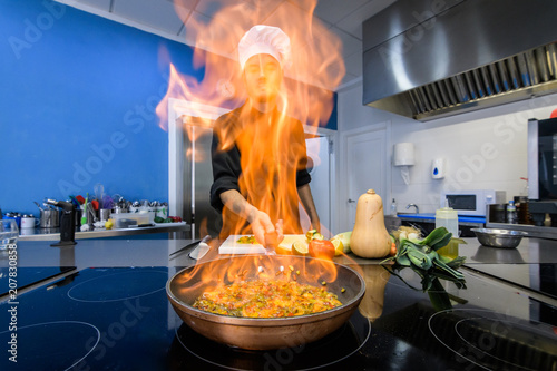 young chef flambe vegetables with fire in a frying pan, front view photo