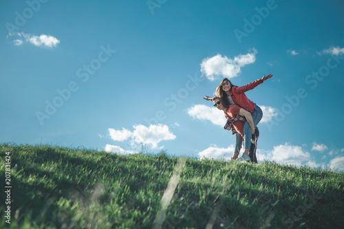 Let us fly. Low angle of smiling young woman and boyfriend fooling around outdoors. Joyful man carrying his girlfriend on back. Excited girl raising hands imitating bird. Copy space in left side