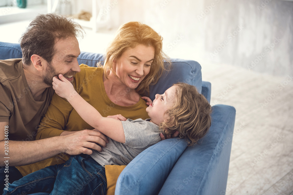 Laughing mom and dad are playing with kid by fondling him. Pleased child is lying on couch near spouse and touching father beard. Happy family concept