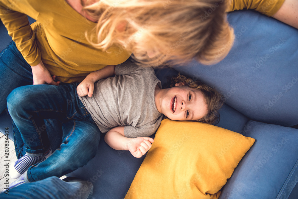 Top view of laughing son spending great time and looking at camera with joy. He is lying on sofa near his mother who is tenderly watching him. Parent is entertaining kid by kindly fondling Top view of laughing son spending great time and looking at camera with joy. He is lying on sofa near his mother who is tenderly watching him. Parent is entertaining kid by kindly fondling