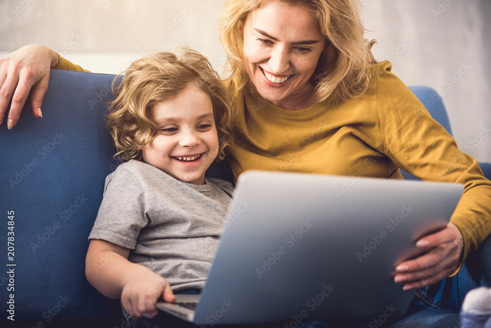 Smiling mom and kid are watching on laptop screen. They are sitting on divan and enjoying time together. Child is holding gadget and laughing while parent is embracing him Smiling mom and kid are watching on laptop screen. They are sitting on divan and enjoying time together. Child is holding gadget and laughing while parent is embracing him