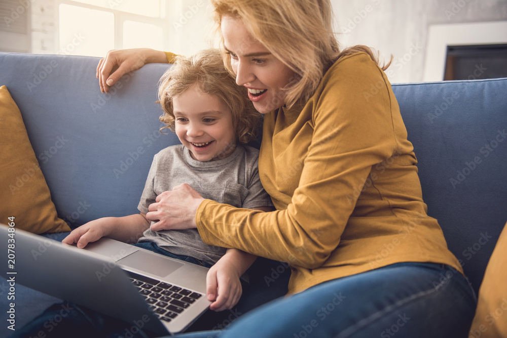Exited mother and little boy are watching something on notebook. They are sitting on sofa and smiling. Woman is hugging kid while he is holding gadget Exited mother and little boy are watching something on notebook. They are sitting on sofa and smiling. Woman is hugging kid while he is holding gadget