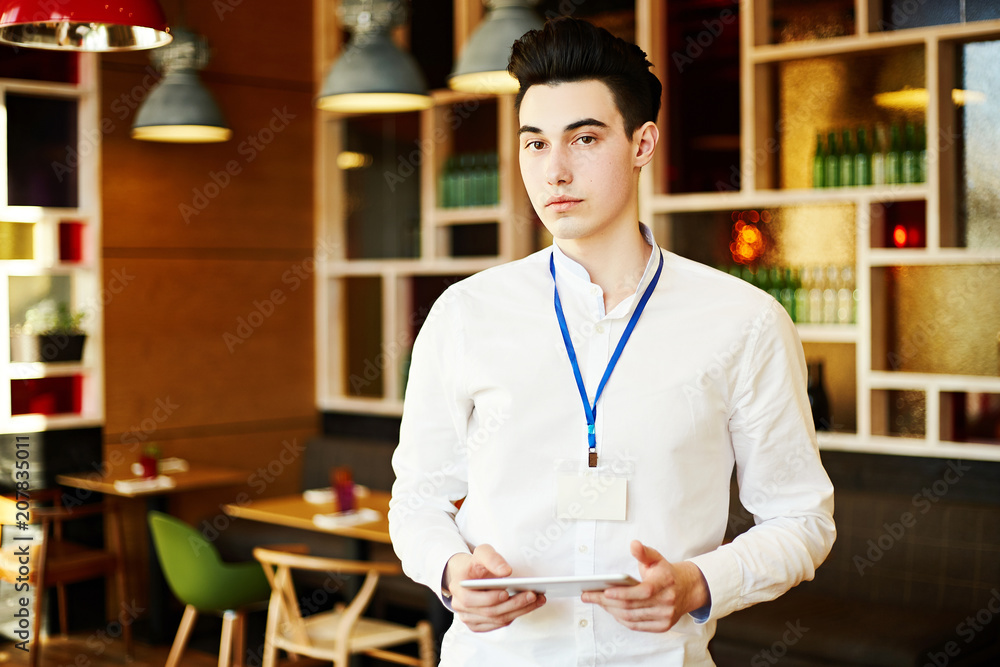 Fototapeta premium Confident young man wearing white shirt and Id card standing with tablet in coffee break room and looking at camera.