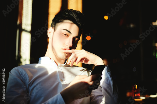 Photography Young serious handsome guy in white shirt touching lips alluringly looking at camera in sunlight and holding phone