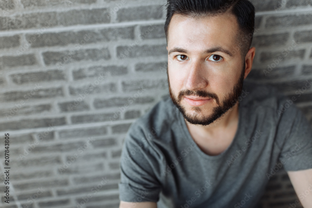 Portrait of a beautiful stylish guy dressed in a grey blank t-shirt standing on a brick grey wall background. Empty space for logo or design. Portrait of a beautiful stylish guy dressed in a grey blank t-shirt standing on a brick grey wall background. Empty space for logo or design.