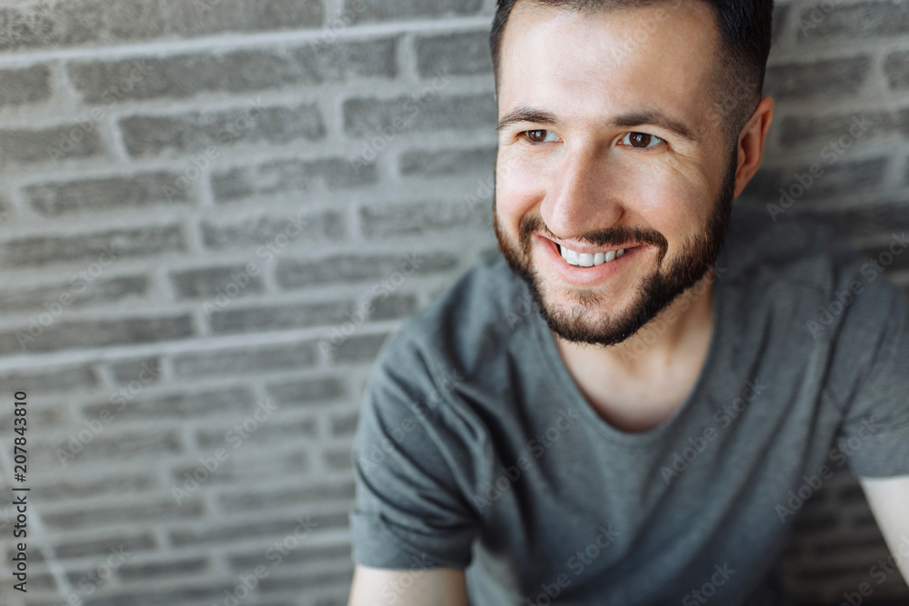 Portrait of a beautiful stylish guy dressed in a grey blank t-shirt standing on a brick grey wall background. Empty space for logo or design. Portrait of a beautiful stylish guy dressed in a grey blank t-shirt standing on a brick grey wall background. Empty space for logo or design.