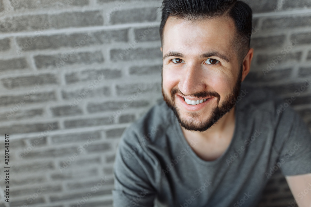 Portrait of a beautiful stylish guy dressed in a grey blank t-shirt standing on a brick grey wall background. Empty space for logo or design. Portrait of a beautiful stylish guy dressed in a grey blank t-shirt standing on a brick grey wall background. Empty space for logo or design.