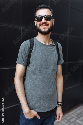 Portrait of a beautiful stylish guy, hipster with glasses, dressed in a gray empty t-shirt, standing on a black wall background. Empty space for logo or design.