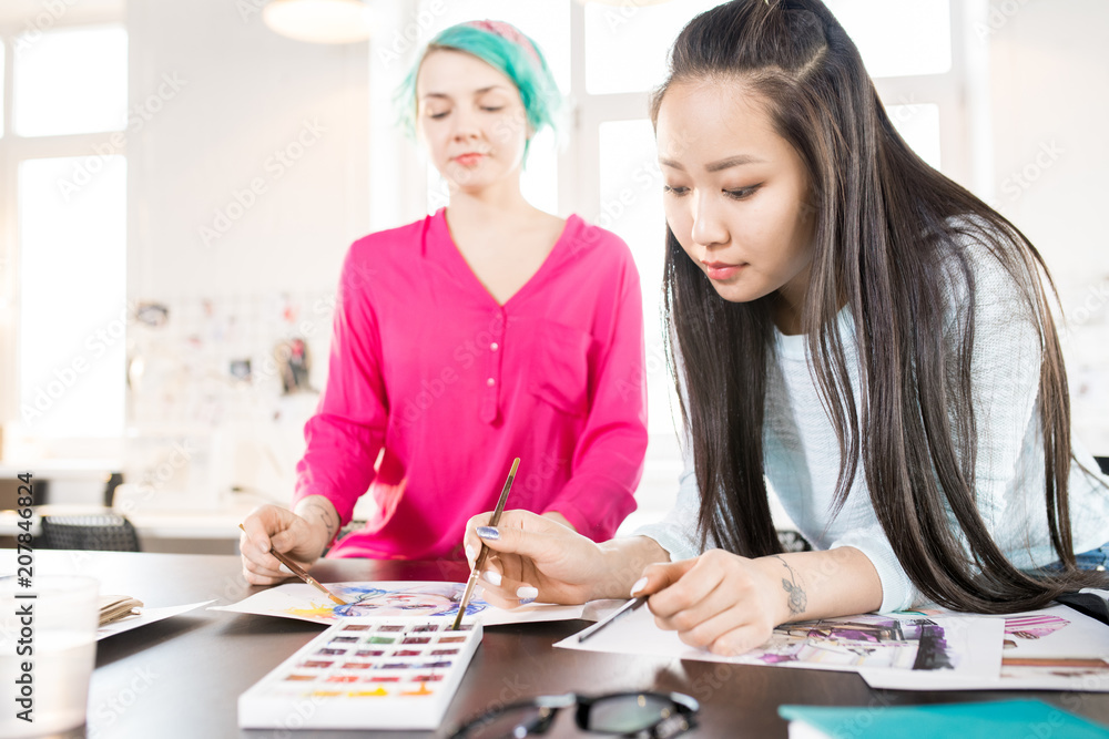 Obraz premium Portrait of two contemporary young women drawing sketches with watercolors on creative design project in small atelier studio, focus on Asian young woman in front, copy space