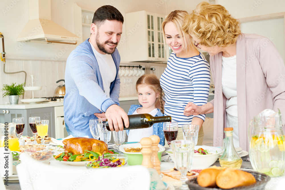 Portrait of happy two generation family standing round festive dinner table together pouring drinks and enjoying delicious homemade dishes during holiday celebration in modern Portrait of happy two generation family standing round festive dinner table together pouring drinks and enjoying delicious homemade dishes during holiday celebration in modern
