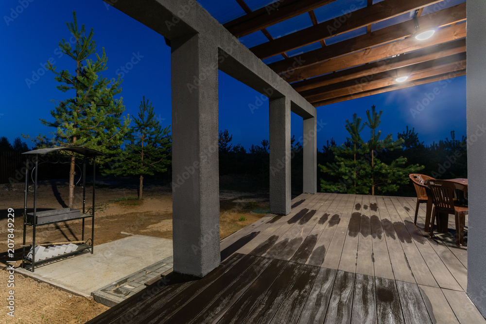 Side view of an open veranda in front of a modern forest cottage. Pine young forest under the sunset rays in the background. Side view of an open veranda in front of a modern forest cottage. Pine young forest under the sunset rays in the background.