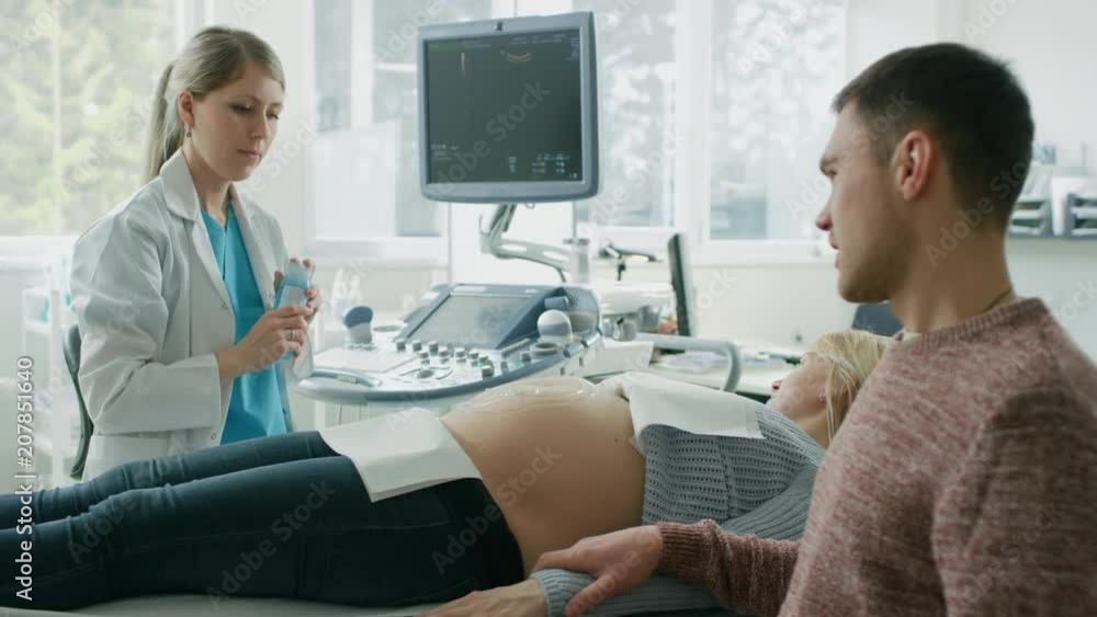 In the Hospital, Female Obstetrician Puts Gel on the Belly of a Pregnant Mother, Ready to do Ultrasound Screening. Husband Diligently Supports His Pregnant Wife. Happy Family. In the Hospital, Female Obstetrician Puts Gel on the Belly of a Pregnant Mother, Ready to do Ultrasound Screening. Husband Diligently Supports His Pregnant Wife. Happy Family.