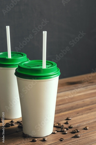 two white plastic coffee cups with a green lid on a wooden table with scattered coffee beans