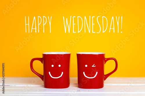 Two red coffee mugs with a smiling faces on a yellow background  with the phrase Happy wednesday.
