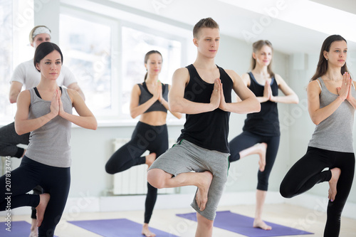 Group of young sporty people practicing yoga lesson, doing Vrksasana exercise, Tree pose, working out, indoor full length, yogi students training in sport club. Wellness, healthy life concept