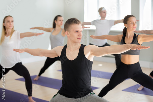 Group of young sporty people practicing yoga lesson, doing Warrior exercise, Virabhadrasana 2 pose, working out, indoor close up, yogi students training in sport club, studio. Active lifestyle concept