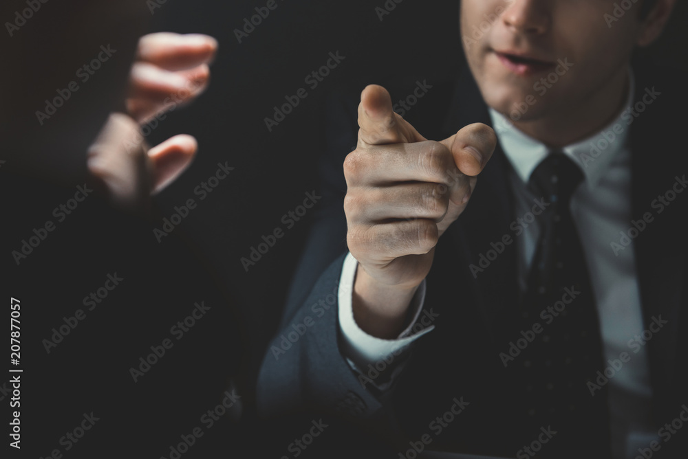 Angry man in formal business suit pointing hand to somesone he talking to in dark interrogation room Angry man in formal business suit pointing hand to somesone he talking to in dark interrogation room