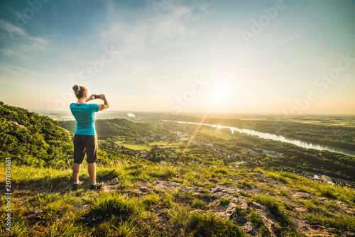 A Girl Standing on the Edge of the Cliff and Taking Photograph of the Sunset and the Valley with the River