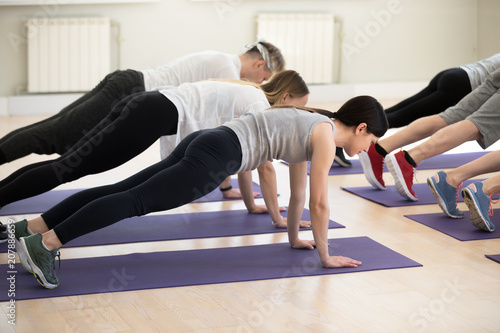 Group of young sporty people practicing yoga lesson, doing Plank exercise, Push ups or press ups, working out, indoor full length, students training in sport club. Active lifestyle, wellness concept