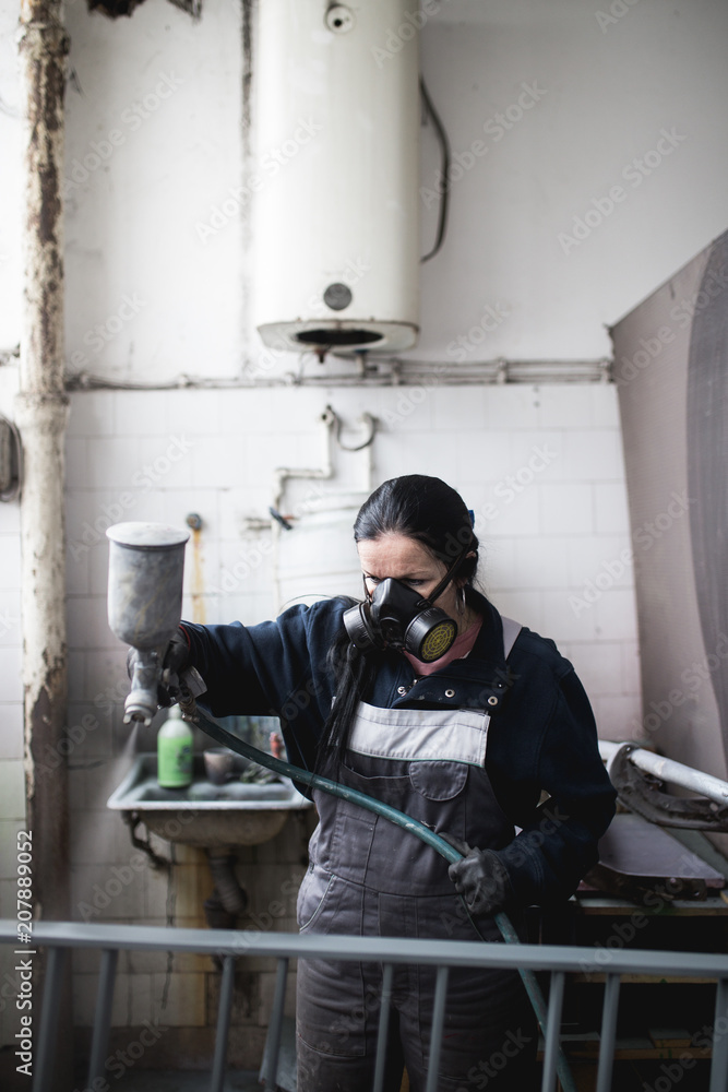 Strong and worthy woman doing hard job. She using industrial spray compressor for painting some metal products.