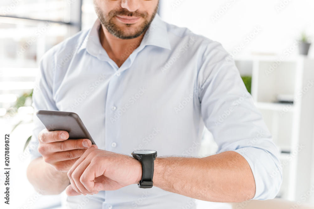 Cropped image closeup of european man in formal wear holding black smartphone and looking at wrist watch while working in bright office room Cropped image closeup of european man in formal wear holding black smartphone and looking at wrist watch while working in bright office room