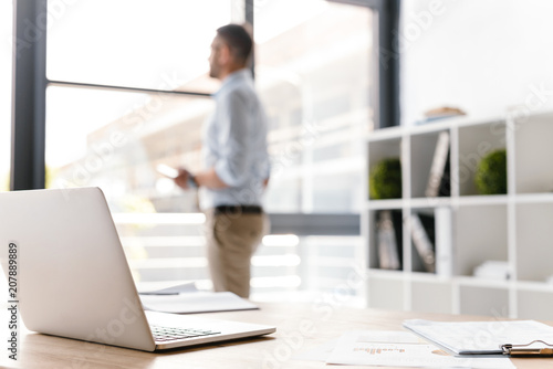 Photo of workplace with open white laptop lying on table, while defocused business man standing and looking through big window on background