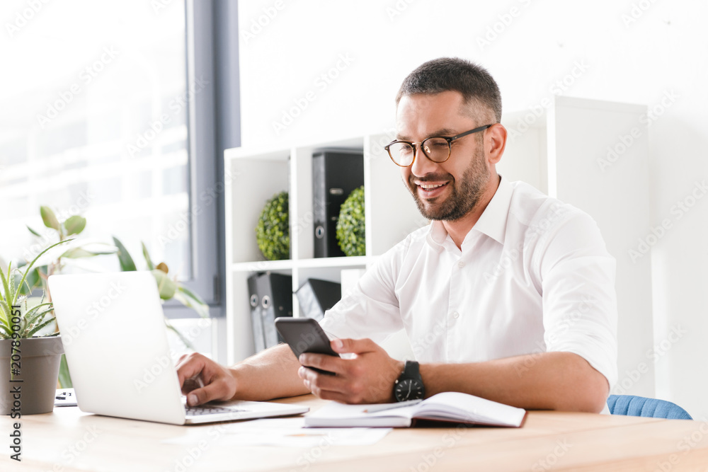Photo of smiling adult man 30s in white shirt using laptop and mobile phone while sitting at table and working in office Photo of smiling adult man 30s in white shirt using laptop and mobile phone while sitting at table and working in office