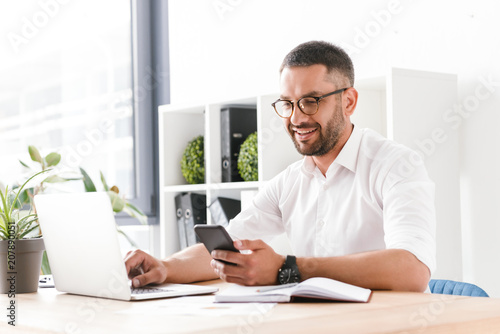 Photo of smiling adult man 30s in white shirt using laptop and mobile phone while sitting at table and working in office