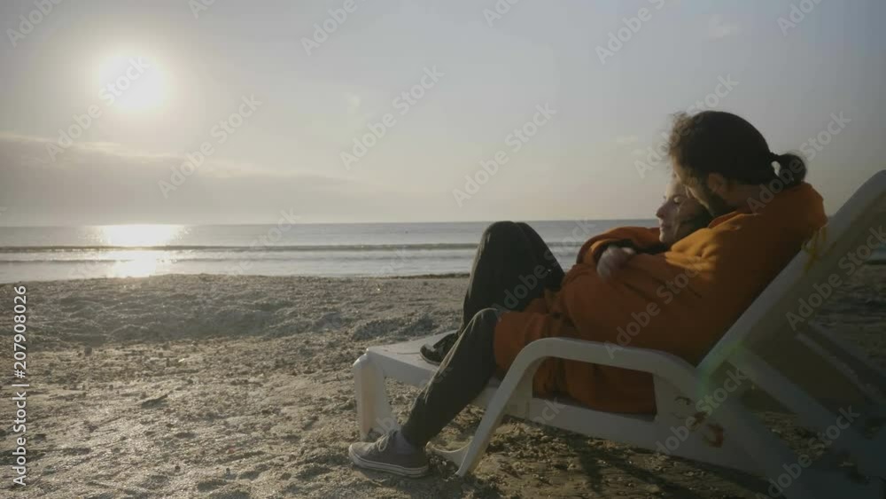 Beautiful young couple covered with a blanket hugging and lying on a lounge chair on the beach enjoying the sunset Beautiful young couple covered with a blanket hugging and lying on a lounge chair on the beach enjoying the sunset